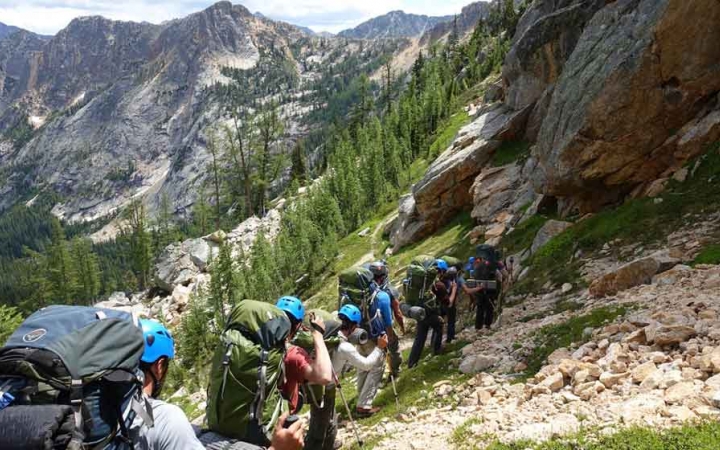 a group of gap year students wearing backpacks and helmets follow a trail up a mountainous landscape